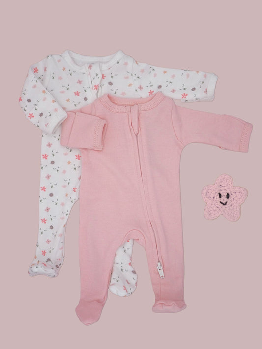 A baby clothing set featuring a pink footed sleeper in the foreground, a white long-sleeve sleeper with a pink and gray floral pattern behind it, and a small pink crocheted star toy on a solid dusty pink background.