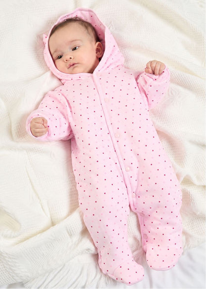 A baby lying on a white textured blanket wearing a pink hooded footed sleeper with small dark polka dots.