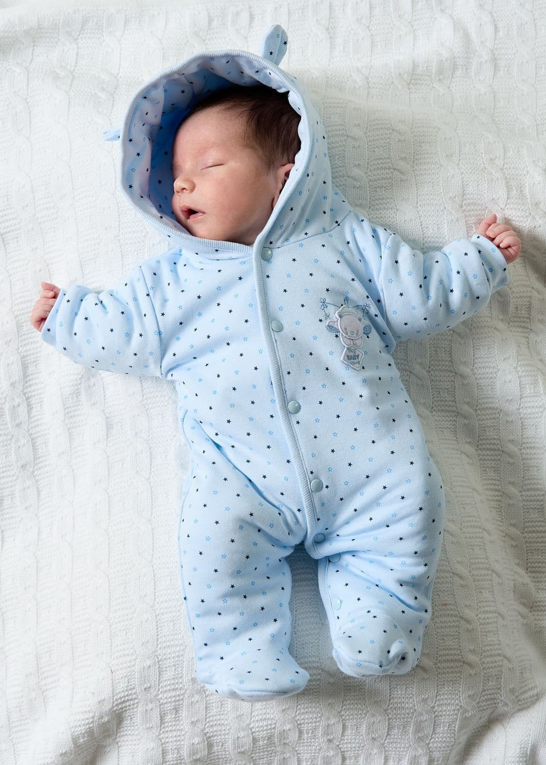 Newborn baby wearing a blue onesie with a hood on a white blanket