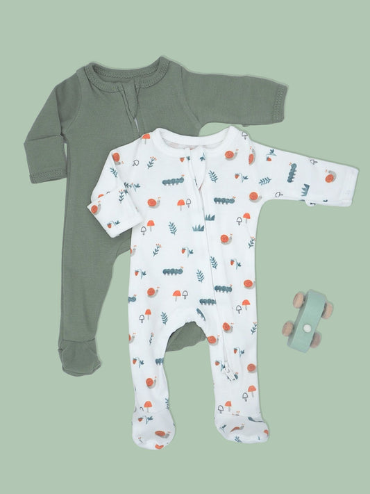 Two baby footed sleepers laid flat on a sage green background: one solid sage green long-sleeve sleeper and one white long-sleeve sleeper with a small multicolor forest and sun pattern. A small toy car sits to the right.