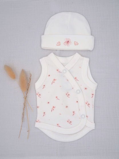 A baby outfit set featuring a white sleeveless wrap bodysuit and matching beanie hat, both decorated with a delicate pink floral pattern, laid flat on a light gray textured background next to dried pampas grass.
