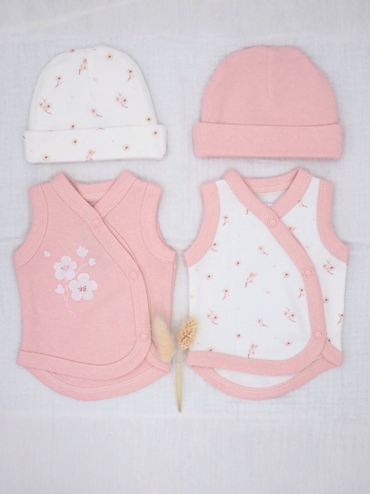 A baby clothing set laid flat on a white textured fabric background, including a pink knit hat, a white knit hat with small pink floral prints, a pink wrap vest, and a white wrap vest with pink trim and tiny pink floral prints. A small dried flower stem lies between the garments.