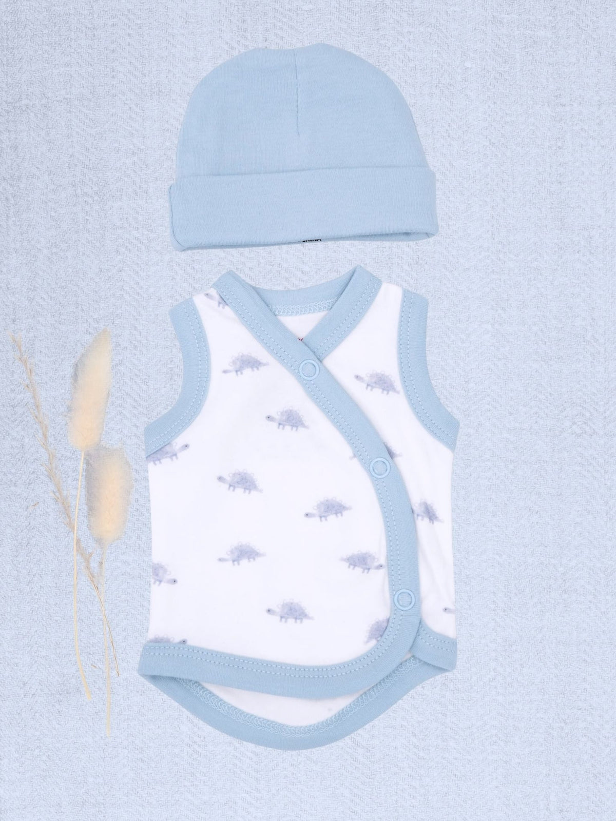 A baby clothing set featuring a white wrap bodysuit with light blue trim and a matching light blue beanie hat, styled flat-lay on a textured light blue background with dried bunny tail grass.