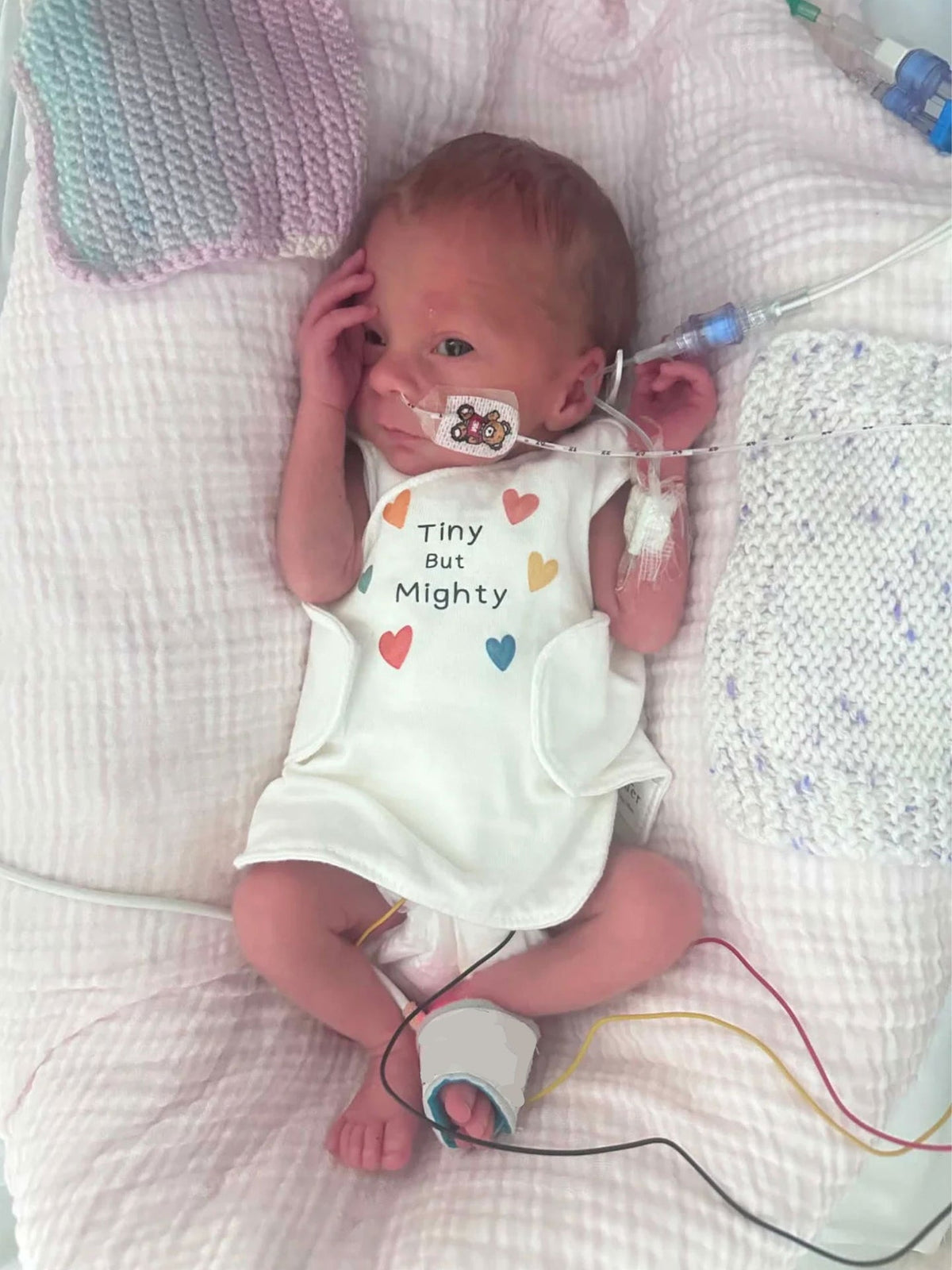 A newborn baby wearing a white sleeveless bodysuit with 'Tiny But Mighty' and colorful heart graphics, lying on a white textured blanket with medical monitoring wires and tubes attached.